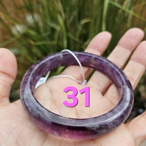 May include: A translucent purple amethyst bangle bracelet. The bracelet is a solid band of polished stone, with variations in color. The number "31" is visible on the bracelet. The background is blurred green foliage.