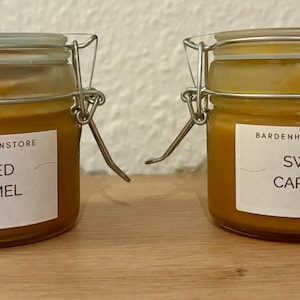 Puede incluir: Dos pequeños tarros de cristal de caramelo con cierres metálicos. Uno está etiquetado como "Salted Caramel" y el otro como "Sweet Caramel". Cada tarro contiene 90 ml de caramelo. Los tarros están sobre una superficie de madera.