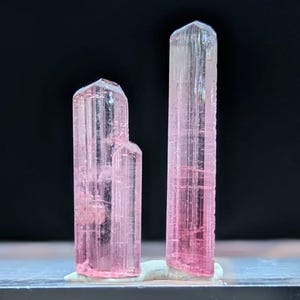 以下が含まれることがあります： 3つのピンク色のトルマリン結晶。結晶は半透明のピンク色で、垂直の縞模様があります。黒い背景に対して、透明な長方形のベースに表示されています。結晶の高さは異なります。