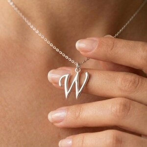 Puede incluir: Un collar plateado con un colgante con forma de la letra "W". El colgante cuelga de una cadena fina. El collar está siendo sostenido por una persona.
