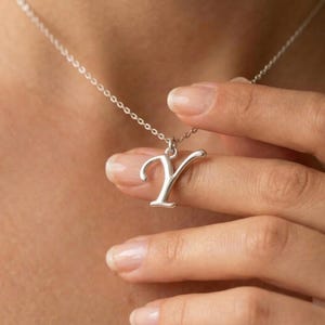 May include: A silver-colored pendant necklace featuring the letter "Y". The pendant is in a cursive font and hangs from a delicate chain. The necklace is being held by a hand, showcasing the jewelry's design and detail.