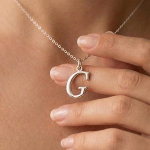 Könnte beinhalten: Eine silberne Halskette mit einem Anhänger in Form des Buchstabens "G". Der Anhänger hängt an einer feinen Kette. Die Halskette wird von einer Hand gehalten, wodurch das Design und die Details des Schmucks hervorgehoben werden.