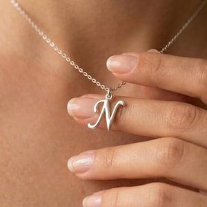 Puede incluir: Un collar plateado con un colgante con la letra "N" en cursiva. El colgante cuelga de una delicada cadena. El collar se muestra sobre un fondo color piel.