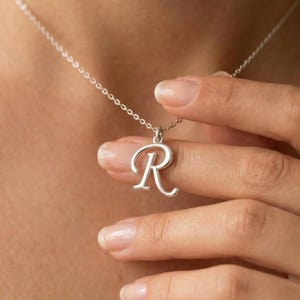 May include: A silver necklace with a cursive letter "R" pendant. The pendant hangs from a delicate chain. The necklace is being held by a hand with manicured nails, showcasing the jewelry's design and detail.