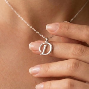 Könnte beinhalten: Eine silberfarbene Anhängerkette mit dem Buchstaben "D". Der Anhänger ist ein Großbuchstabe in einer schlichten, eleganten Schriftart. Die Halskette ist filigran und silberfarben. Die Halskette wird von einer Person gehalten.