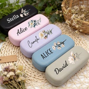 Pode incluir: Cinco estojos de óculos personalizados em preto, rosa, lavanda, azul e cinza. Cada estojo apresenta um nome em escrita cursiva e um design floral. Os estojos estão dispostos sobre uma esteira tecida com um pequeno buquê e uma cesta ao fundo.