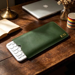 May include: A dark green rectangular sleeve case with the gold embossed initials ES on the bottom right corner. The sleeve is partially open, revealing a white wireless keyboard with rounded keys protruding from the left side of the case.