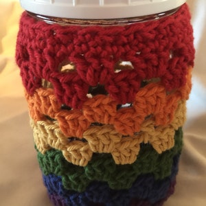 May include: A glass jar with a white lid is covered in a rainbow crochet pattern. The crochet is made in red, orange, yellow, green, blue, and purple yarn.