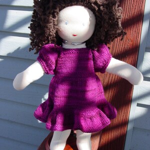May include: A handmade doll with brown curly hair, wearing a purple knitted dress. The doll has a white face with painted blue eyes and a pink mouth.