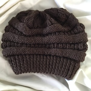 May include: A brown knitted beanie hat with a slouchy, folded design. The hat is made of a soft, chunky yarn and has a ribbed texture.