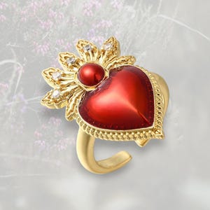 May include: A gold-toned ring featuring a vibrant red heart-shaped centerpiece. The heart is surrounded by a gold frame with a decorative edge. The ring is accented with a sunburst design and small, sparkling gemstones.
