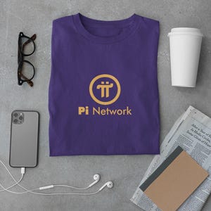 May include: A purple t-shirt with a gold Pi Network logo and text. The t-shirt is folded and laid flat on a gray surface. There is a smartphone, a pair of eyeglasses, a cup of coffee, a notebook, and a pair of earbuds in the image.