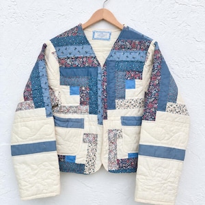 May include: A long-sleeve quilted jacket featuring a patchwork design of blue, white, and floral fabric squares. The jacket has a V-neckline, a front buttonless opening, and two front pockets, all constructed from stitched fabric segments.