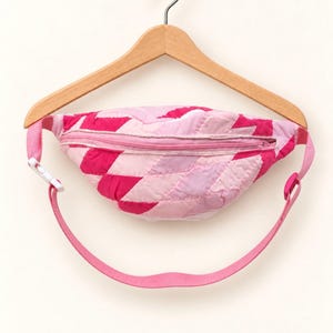 May include: A quilted fanny pack featuring a geometric pattern in shades of light pink, medium pink, and hot pink. The bag has a horizontal zippered closure across the top and an adjustable pink strap with a white plastic side-release buckle.