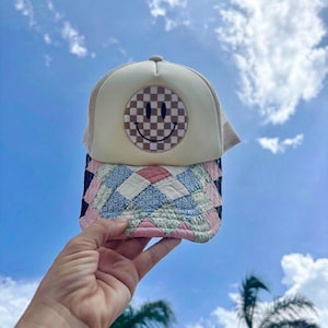 May include: A trucker hat featuring a cream-colored front panel with a brown and white checkered smiley face patch. The brim is covered in a quilted fabric pattern consisting of diamond-shaped patches in shades of pink, blue, and green floral prints.