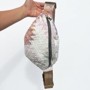 May include: A quilted fanny pack featuring a patchwork design with floral and geometric patterns in shades of white, pink, and grey. The bag includes a dark grey vertical zipper closure and an adjustable tan nylon strap with a plastic buckle.