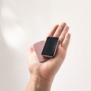May include: A small, rectangular, dusty rose-colored metal tin with a matching lid. The tin features rounded corners and a smooth, matte finish. The lid is slightly offset, revealing a solid black interior surface within the container.