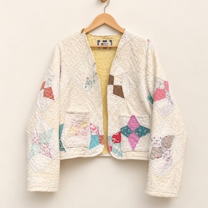 May include: A long-sleeved quilted jacket made from cream-colored fabric with a patchwork pattern of multi-colored floral and geometric shapes. The jacket features an open front, two front patch pockets, and a solid yellow interior lining.
