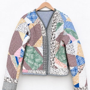 May include: A long-sleeve quilted jacket constructed from various patchwork fabric scraps in shades of blue, green, pink, yellow, and white. The garment features a V-neckline, a gray trim along the edges, and a small blue and white checkered lining.