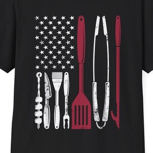 May include: A black short-sleeved t-shirt featuring a graphic print of an American flag design composed of white stars and red barbecue tools, including a skewer, knife, brush, fork, spatula, tongs, and a bottle opener arranged in vertical stripes.