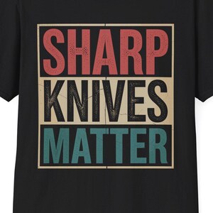Peut inclure: T-shirt noir avec l'inscription "SHARP KNIVES MATTER" dans un style rétro. Le texte est disposé sur trois lignes : "SHARP" en rouge, "KNIVES" en beige et "MATTER" en bleu sarcelle. Col rond classique.