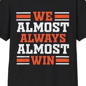 May include: Black t-shirt with the text "WE ALMOST ALWAYS ALMOST WIN" in white and orange block letters. The shirt has a crew neck and short sleeves. The text is centered on the front of the shirt.