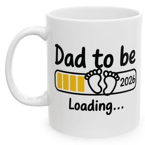May include: White ceramic mug with the text "Dad to be Loading... 2026". The design includes a loading bar and baby footprints in black and yellow. The mug has a curved handle.