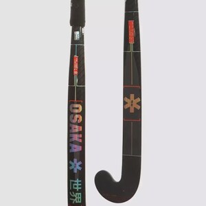 Op de afbeelding: Twee zwarte veldhockeysticks met koolstofvezeltextuur. De linkerstick toont de tekst OSAKA en Japanse karakters in een regenboogverloop, met een ster-symbool. De rechterstick toont een ster-icoon en rode rechthoekige labels.