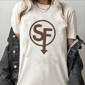 Op de afbeelding: Crèmekleurig T-shirt met een bruine cirkelvormige afbeelding met de letters "SF" en een pijl naar beneden. Het shirt is gemaakt van zacht materiaal en heeft korte mouwen. Een donker spijkerjasje is over de schouders gedrapeerd.