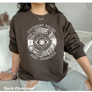 Puede incluir: Sudadera de color chocolate oscuro con un dise&ntilde;o gr&aacute;fico blanco. El dise&ntilde;o presenta un s&iacute;mbolo de ojo central rodeado por un patr&oacute;n circular con las palabras "STATEMENT BEGINS" y "STATEMENT ENDS". La sudadera tiene mangas largas y cuello redondo.