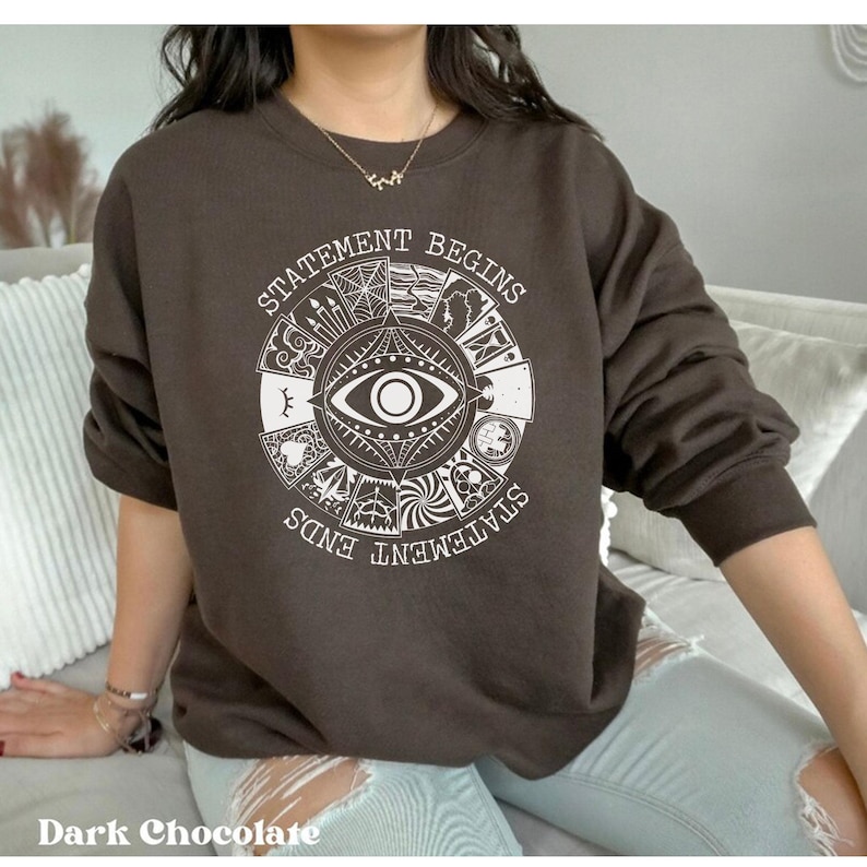 Puede incluir: Sudadera de color chocolate oscuro con un dise&ntilde;o gr&aacute;fico blanco. El dise&ntilde;o presenta un s&iacute;mbolo de ojo central rodeado por un patr&oacute;n circular con las palabras "STATEMENT BEGINS" y "STATEMENT ENDS". La sudadera tiene mangas largas y cuello redondo.