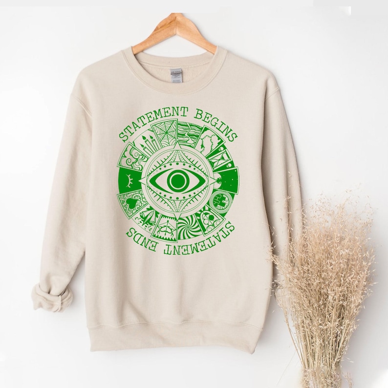 Puede incluir: Sudadera beige con un dise&ntilde;o gr&aacute;fico verde. El dise&ntilde;o presenta un ojo en el centro, rodeado de varios s&iacute;mbolos y las palabras "STATEMENT BEGINS" y "STATEMENT ENDS" en un patr&oacute;n circular. La sudadera est&aacute; colgada en una percha de madera.
