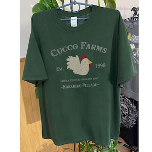 May include: Dark green t-shirt with a distressed graphic that reads "CUCCO FARMS" above a rooster illustration. The text "EST. 1998" and "ATTACK CUCCO AT YOUR OWN RISK - KAKARIKO VILLAGE-" are also printed on the shirt.