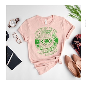 Puede incluir: Una camiseta rosa claro con un dise&ntilde;o gr&aacute;fico verde que presenta un ojo y el texto "STATEMENT BEGINS STATEMENT ENDS". La camiseta est&aacute; combinada con un reloj, gafas, un cuaderno y zapatos.