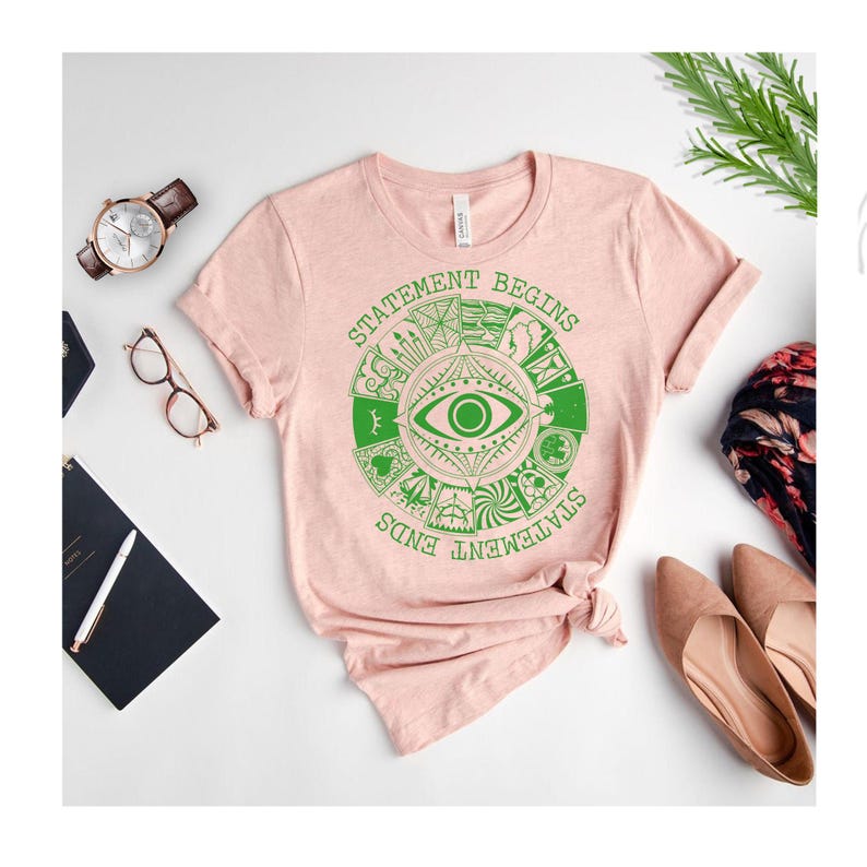 Puede incluir: Una camiseta rosa claro con un dise&ntilde;o gr&aacute;fico verde que presenta un ojo y el texto "STATEMENT BEGINS STATEMENT ENDS". La camiseta est&aacute; combinada con un reloj, gafas, un cuaderno y zapatos.