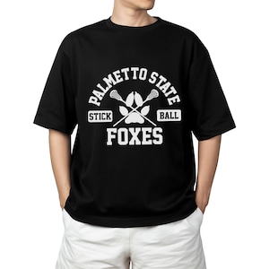 Pu&ograve; includere: T-shirt nera con grafica bianca. La grafica include il testo "PALMETTO STATE" sopra bastoni da lacrosse incrociati e un'impronta di zampa. Sotto, le parole "STICK", "BALL" e "FOXES".