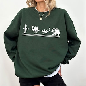 Pode incluir: Camisola verde escuro com um gráfico branco de quatro animais estilizados. Os animais são um coelho, um camelo, um lagarto e um elefante. A camisola tem gola redonda e mangas compridas.