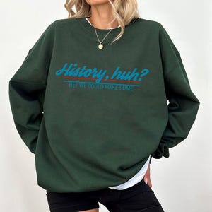 Può includere: Una felpa verde scuro con la scritta "History, huh? Bet we could make some" in blu e rosso. La felpa ha un girocollo e maniche lunghe. La modella indossa pantaloncini neri e una maglietta bianca.