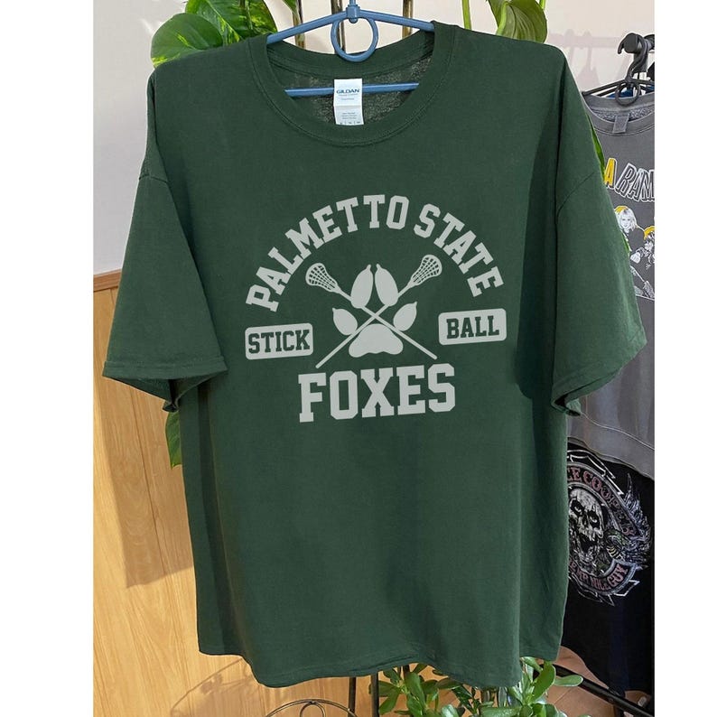 Pu&ograve; includere: Una maglietta verde scuro con la scritta bianca "PALMETTO STATE STICK BALL FOXES". Il design include bastoni da lacrosse e un'impronta di zampa. La maglietta &egrave; appesa a un gancio.