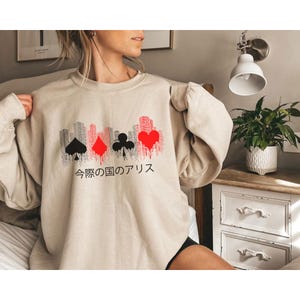 Puede incluir: Sudadera beige con un dise&ntilde;o de paisaje urbano con s&iacute;mbolos de naipes en negro y rojo. El texto japon&eacute;s "Alicia en el pa&iacute;s de las maravillas" est&aacute; impreso debajo. La sudadera tiene cuello redondo y mangas largas.