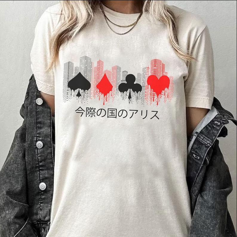 Puede incluir: Camiseta color crema con un dise&ntilde;o de paisaje urbano con palos de cartas rojas y negras. El texto japon&eacute;s "Alice in Borderland" est&aacute; impreso debajo. La camiseta est&aacute; hecha de un material suave.