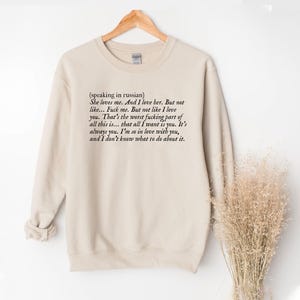 May include: A beige sweatshirt with the text "(speaking in russian) She loves me. And I love her. But not like... Fuck me. But not like I love you..." printed in black. The sweatshirt is hanging on a wooden hanger.