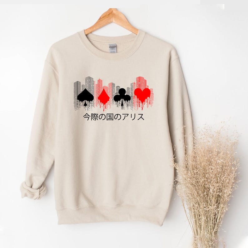 Puede incluir: Sudadera beige con un dise&ntilde;o gr&aacute;fico que presenta una silueta de ciudad y palos de cartas en negro y rojo. Texto japon&eacute;s impreso debajo del dise&ntilde;o. La sudadera est&aacute; colgada en una percha de madera.