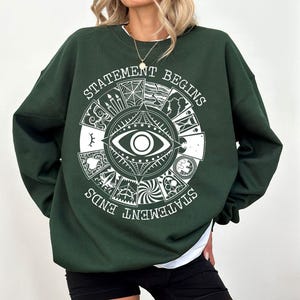 Puede incluir: Sudadera verde oscuro con un diseño gráfico blanco que presenta un ojo, imágenes de tarot y el texto "STATEMENT BEGINS STATEMENT ENDS". La sudadera tiene mangas largas y cuello redondo.