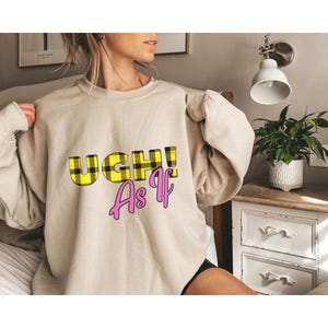 Op de afbeelding: Beige sweatshirt met de tekst "UGH! As If". Het woord "UGH!" is in een geel en zwart geruit patroon, en "As If" is roze.