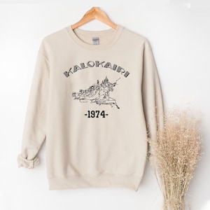 Op de afbeelding: Beige sweatshirt met het woord "KALOKAIRI" boven een zwart-wit illustratie van een gebouw en het jaartal "-1974-". De sweatshirt hangt aan een houten hanger.