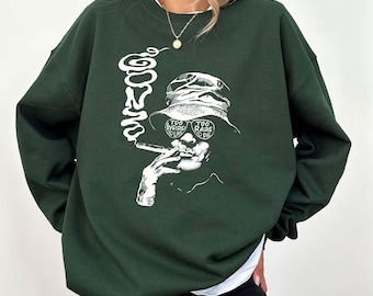 May include: Dark green sweatshirt featuring a white graphic of a person smoking a cigarette, wearing a bucket hat with the text "Too Weird to Live, Too Rare to Die." The sweatshirt has a crew neck and long sleeves.