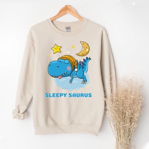 May include: Beige sweatshirt featuring a blue dinosaur wearing a nightcap, resting on a cloud with a crescent moon and star. The text "SLEEPY SAURUS" is printed in blue below the dinosaur. The sweatshirt is hanging on a wooden hanger.