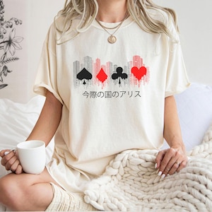 Puede incluir: Camiseta color crema con un dise&ntilde;o gr&aacute;fico de un paisaje urbano y palos de cartas en negro y rojo. Texto japon&eacute;s debajo del dise&ntilde;o. La camiseta tiene mangas cortas y un corte holgado.