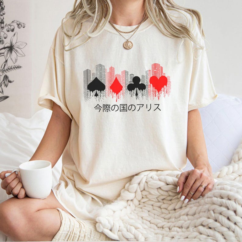 Puede incluir: Camiseta color crema con un dise&ntilde;o gr&aacute;fico de un paisaje urbano y palos de cartas en negro y rojo. Texto japon&eacute;s debajo del dise&ntilde;o. La camiseta tiene mangas cortas y un corte holgado.